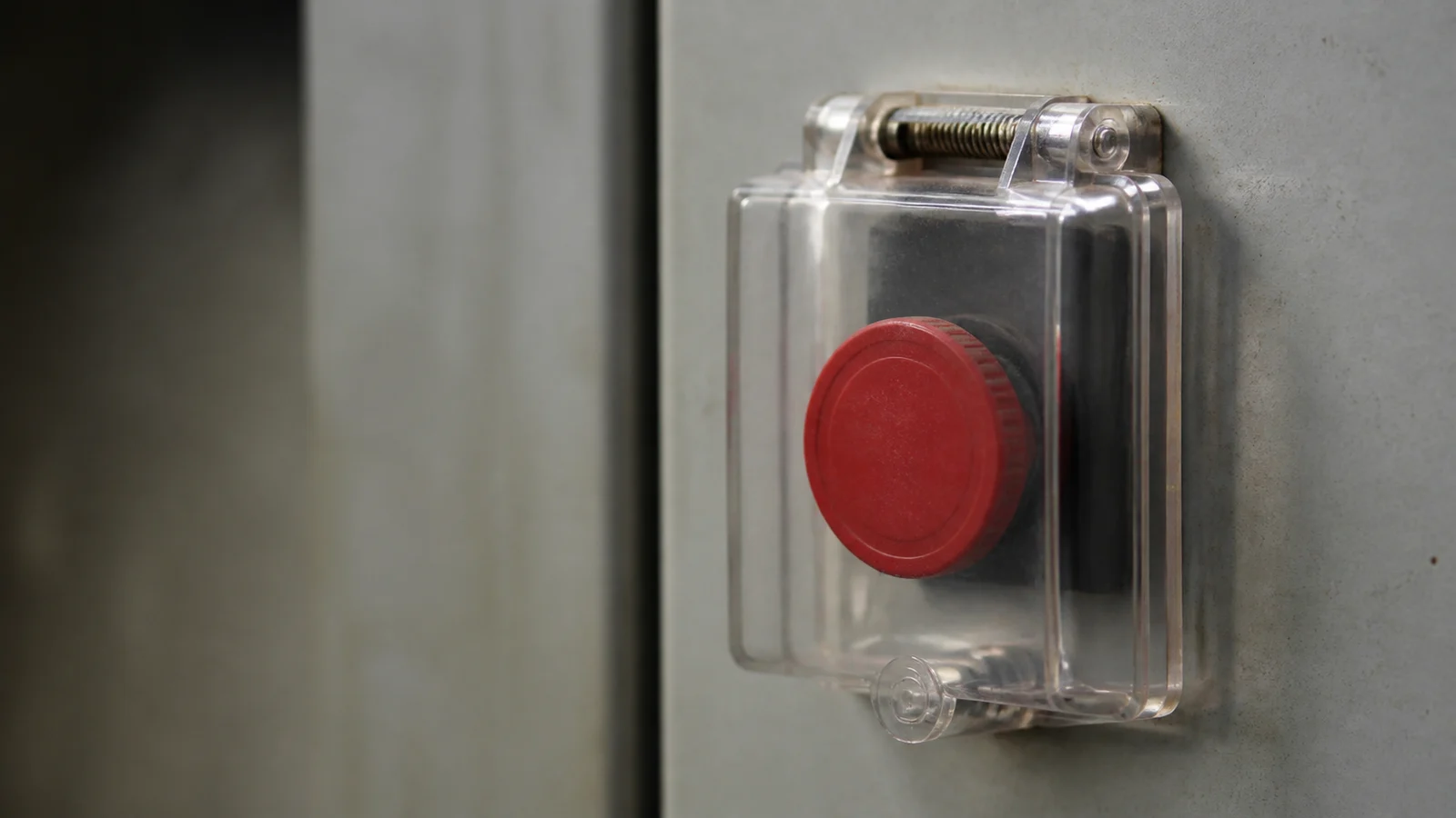 A red industrial push button beneath a closed transparent safety cover on a worn grey control panel
