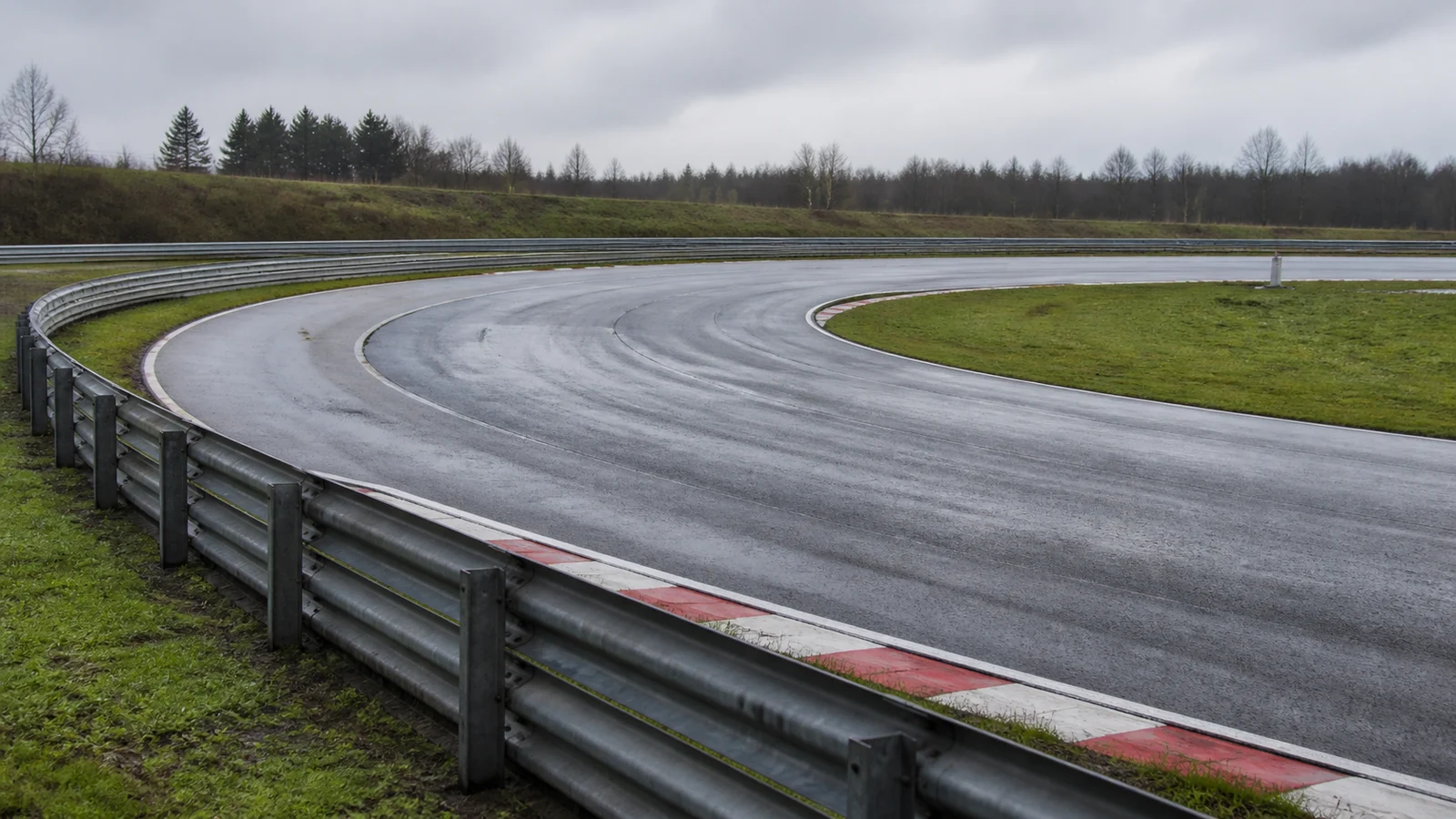 An empty rain-darkened test track curving between metal safety barriers beneath an overcast sky